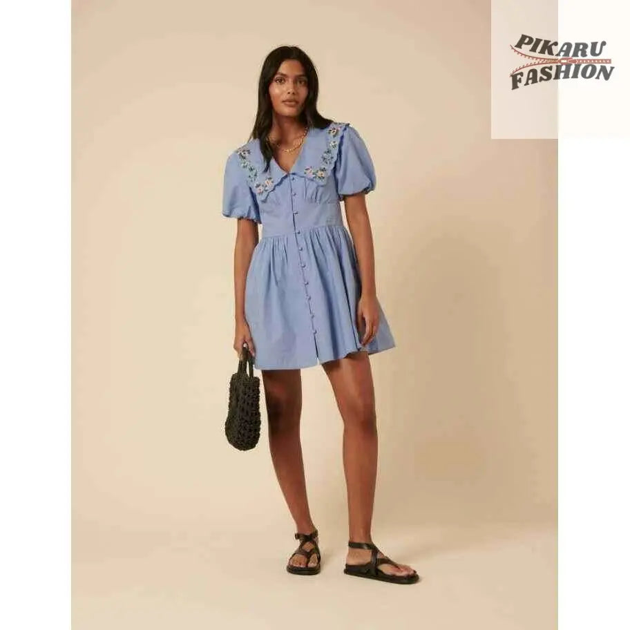 Woman wearing blue puff sleeve mini dress with floral embroidered collar, holding black bag