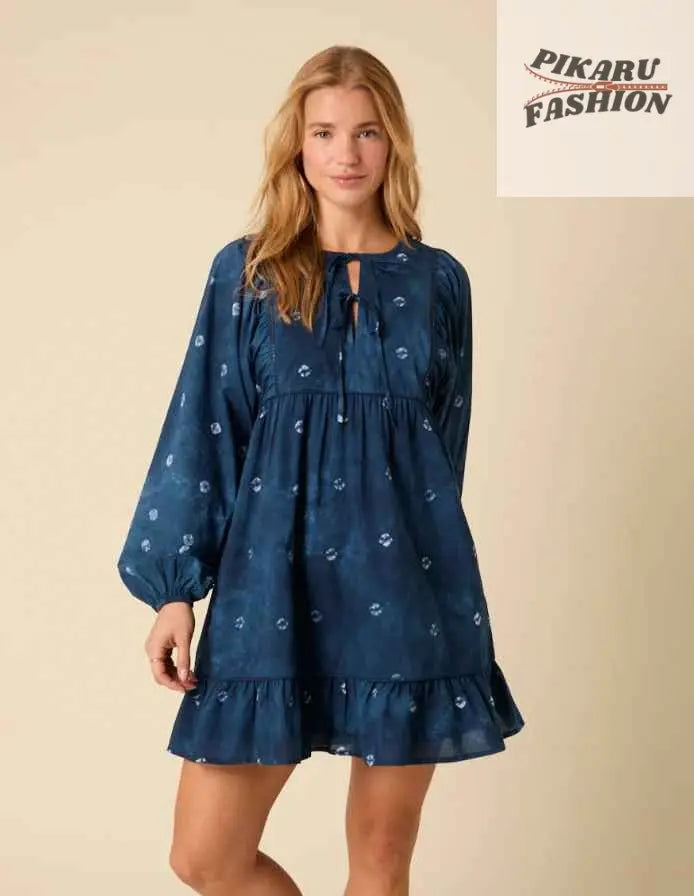 Woman in a blue long-sleeve floral mini dress with ruffle hem standing against beige background