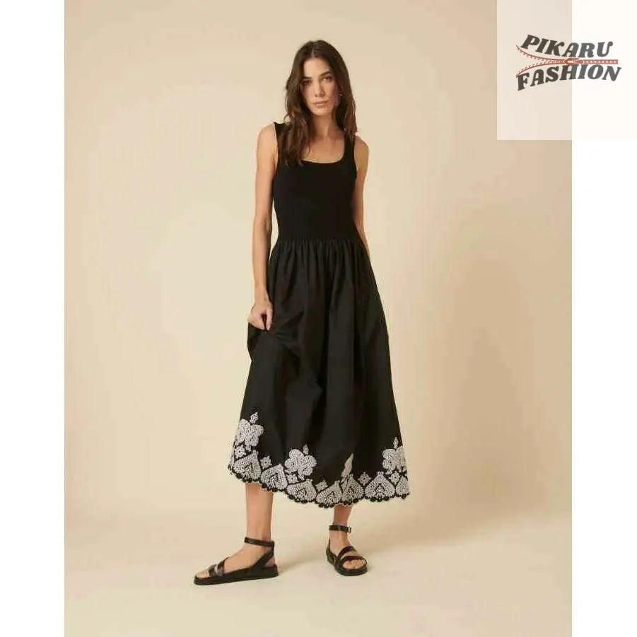 Woman wearing black sleeveless midi dress with white floral hem embroidery, sandals, studio setting