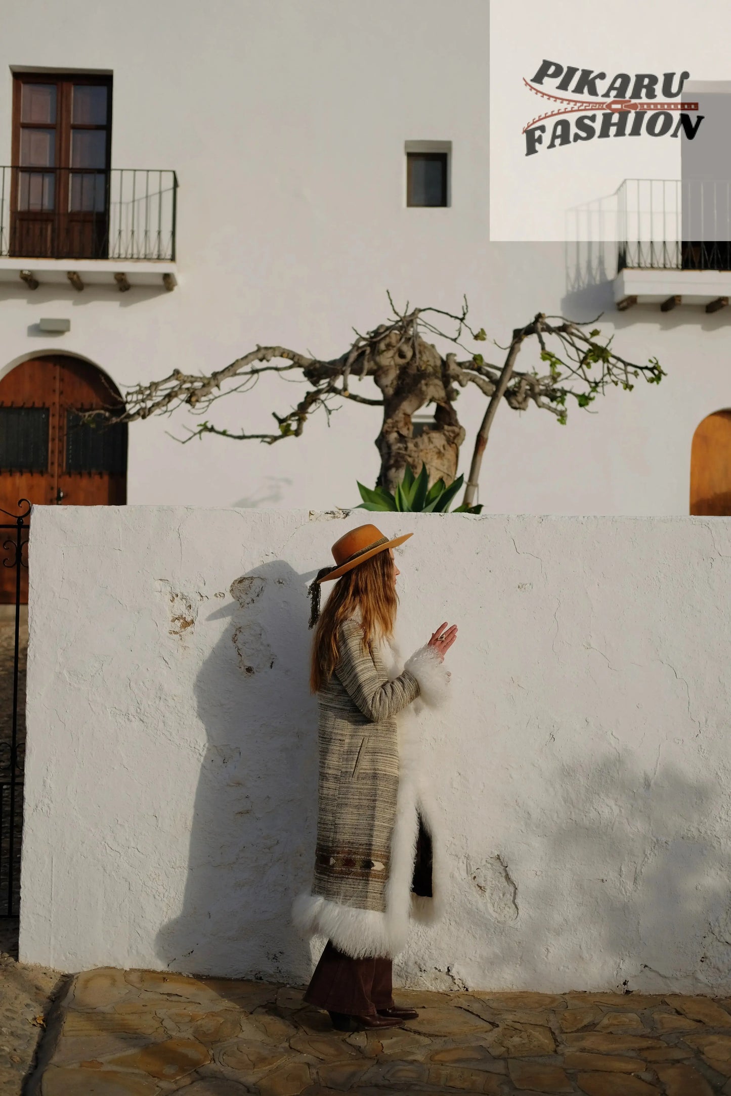 Woman wearing a boho fur trim long coat and brown hat standing outdoors beside a white wall