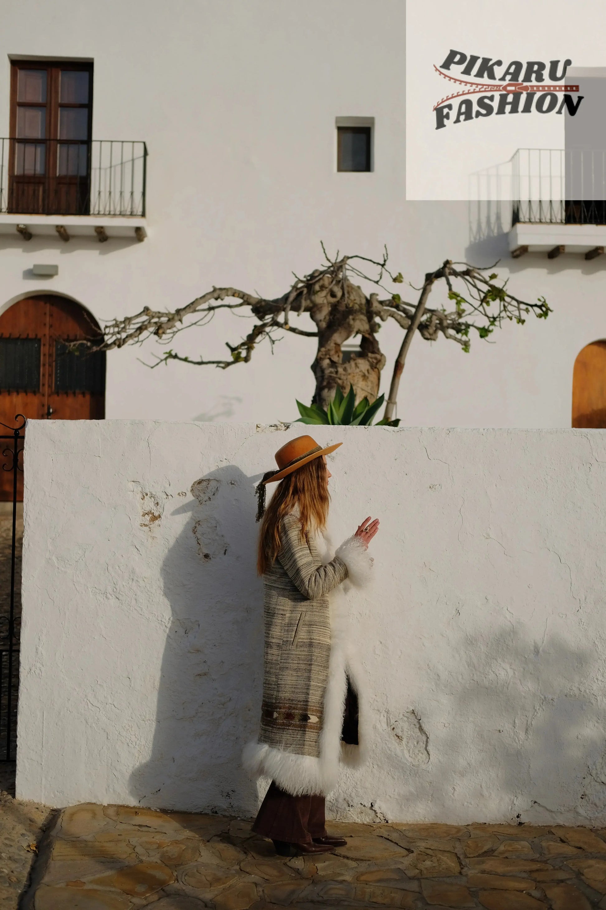Woman wearing a boho fur trim long coat and brown hat standing outdoors beside a white wall
